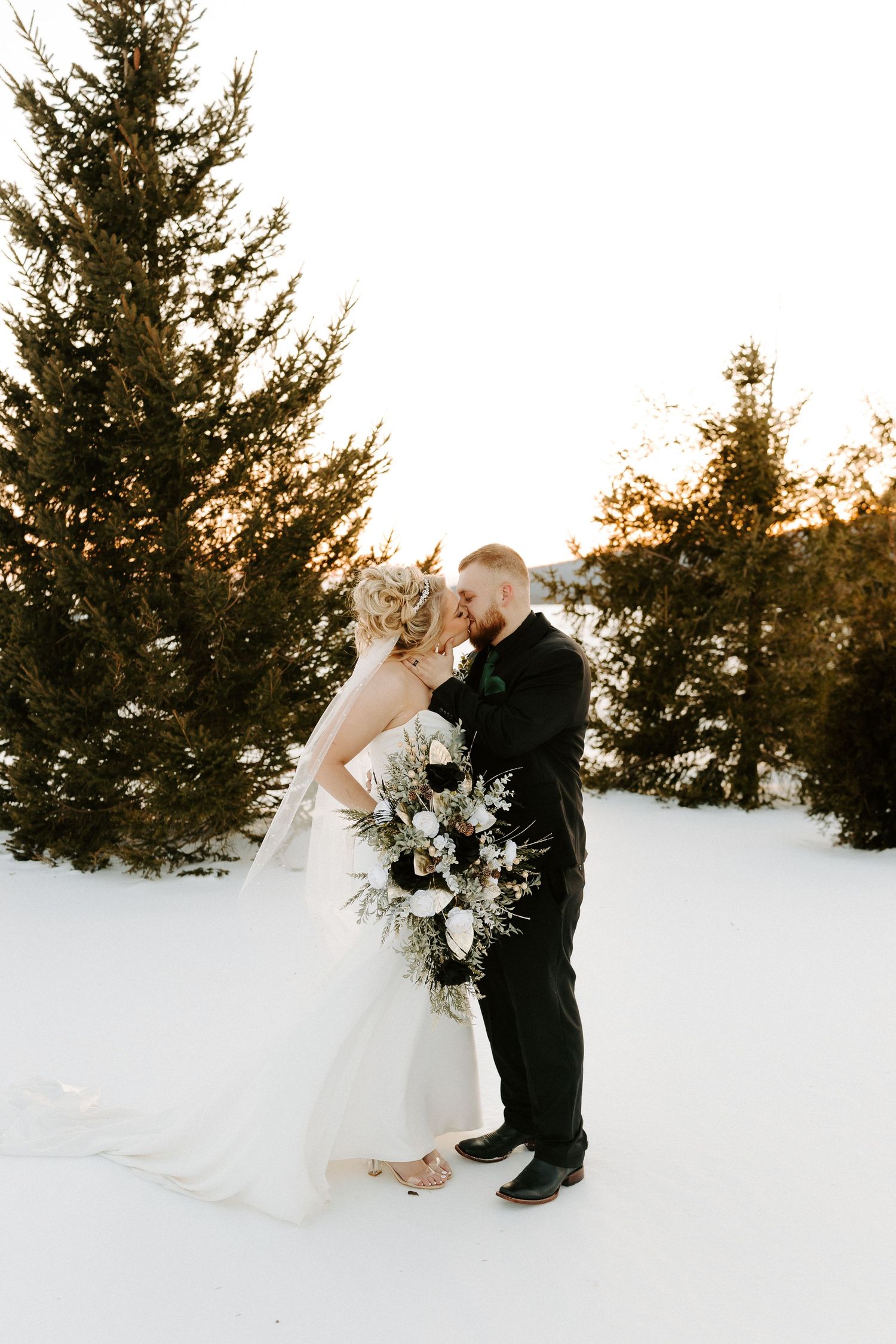 Bride and groom kiss in snowy field surrounded by pine trees at golden hour winter wedding.