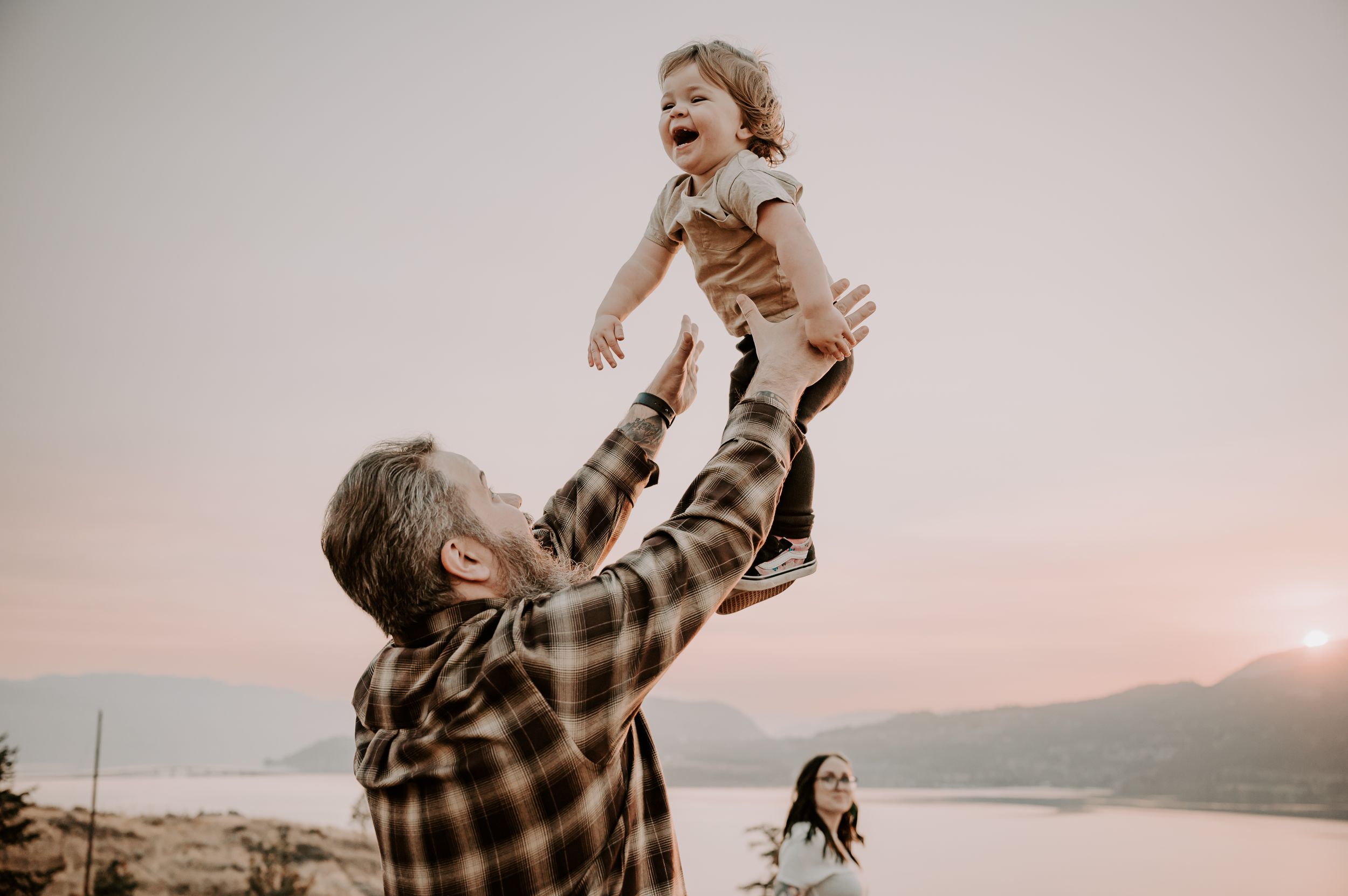 A person lifts a joyful child up high against a scenic mountain landscape at sunset while another group plays in background.