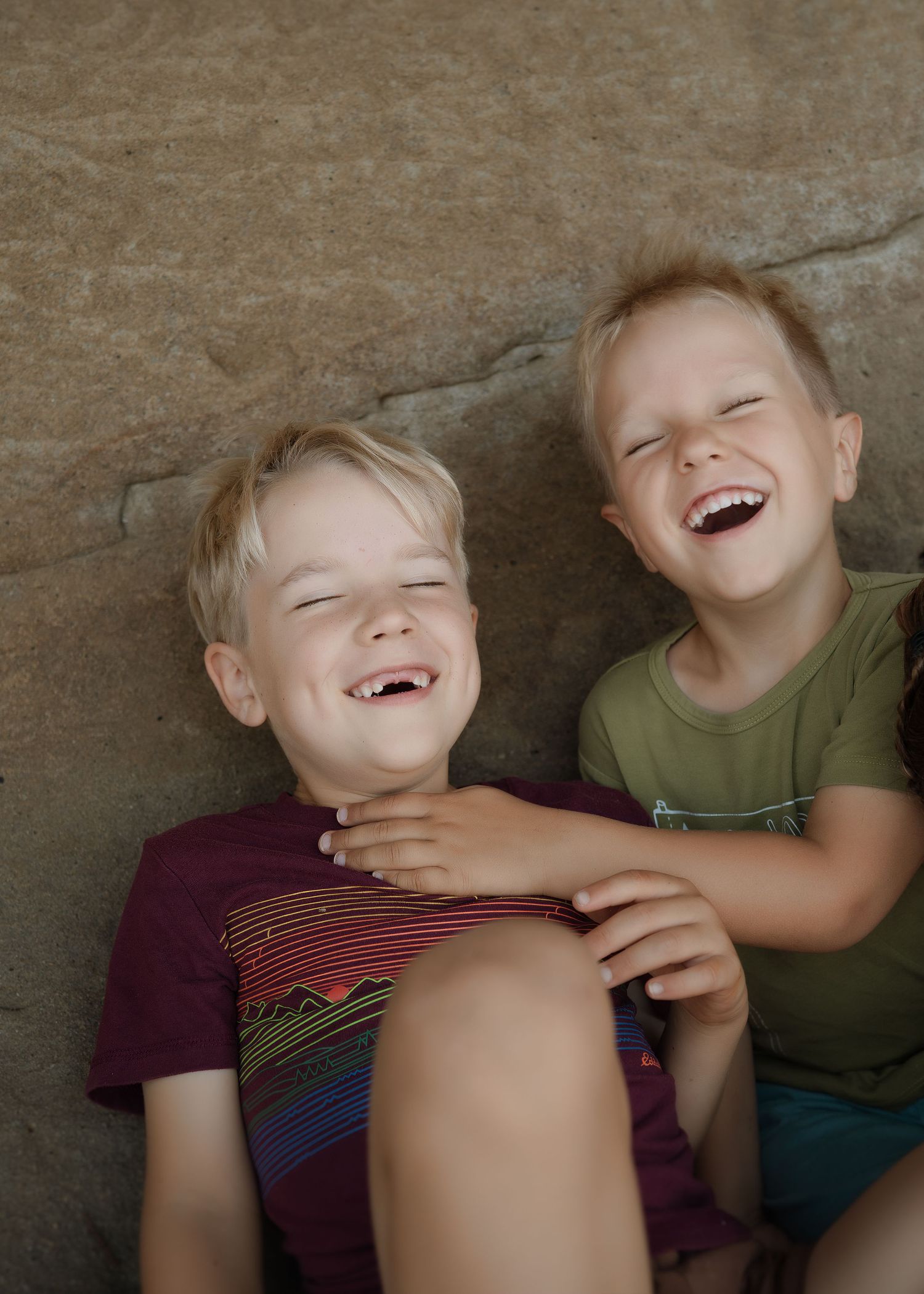 Two young boys laughing joyfully together against a rock wall, one in a maroon shirt and the other in an olive green shirt.