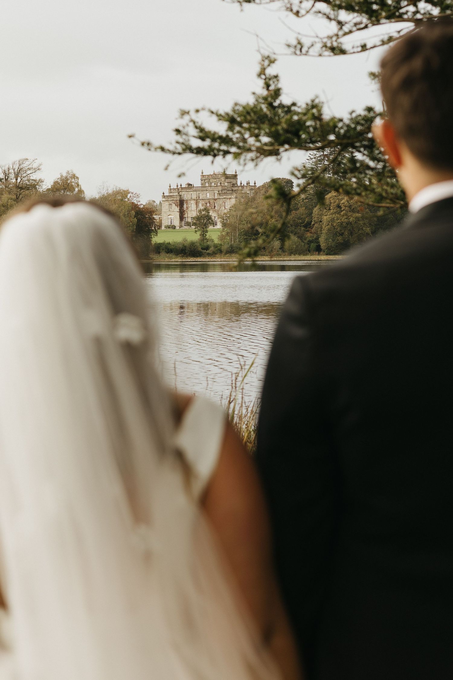 Newlywed couple gather by a tranquil lake with a historic castle visible in the background on an autumn day.