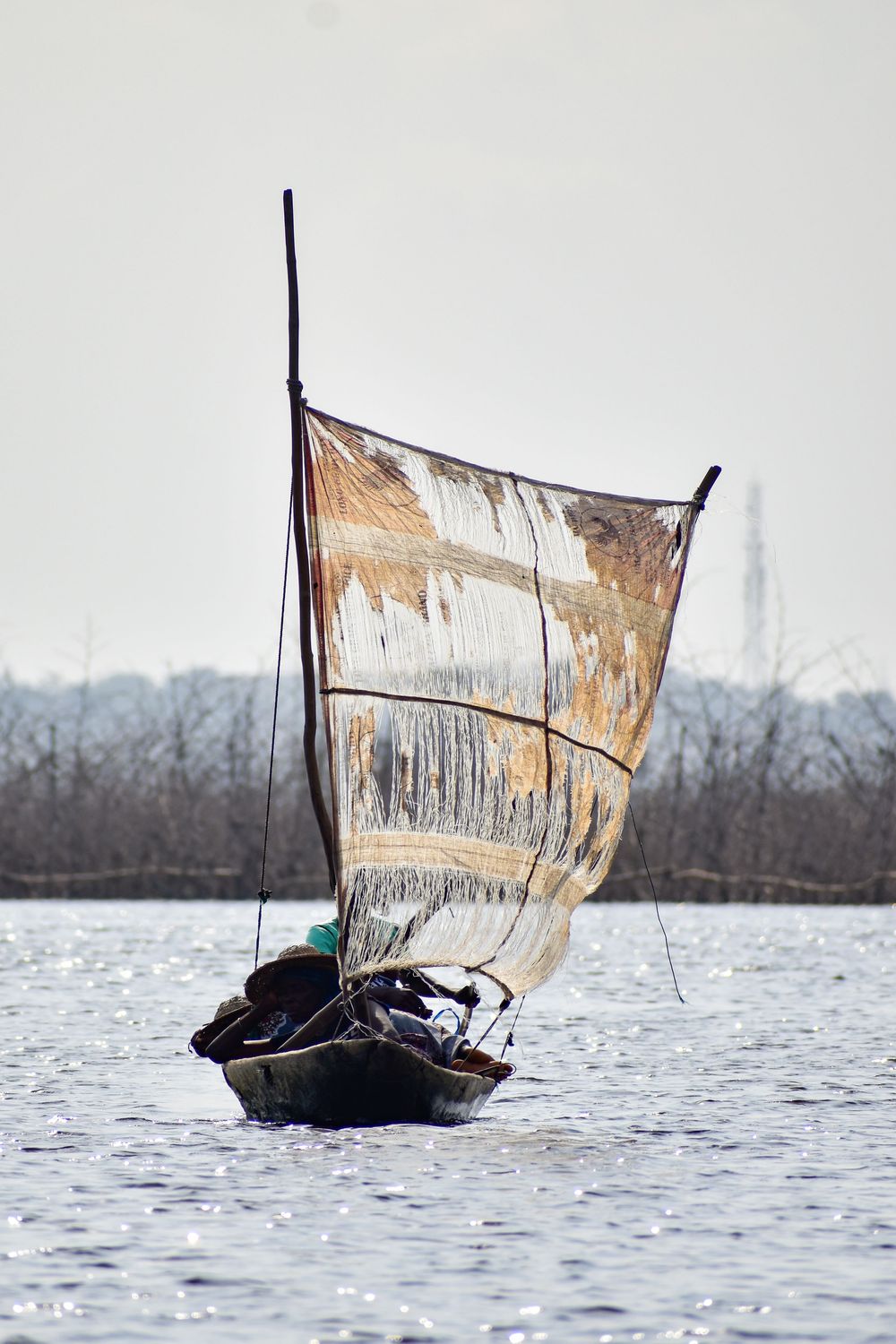 A traditional sailboat with weathered canvas sail navigates across a misty lake during winter.