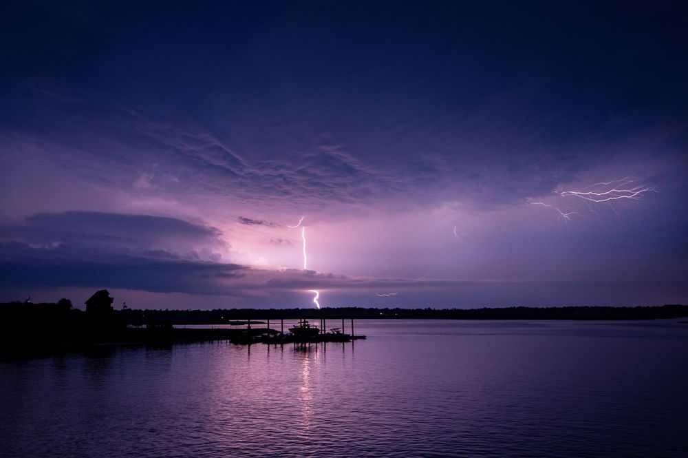 Lightning strikes illuminate the dark sky over a tranquil lake during a dramatic thunderstorm at night.