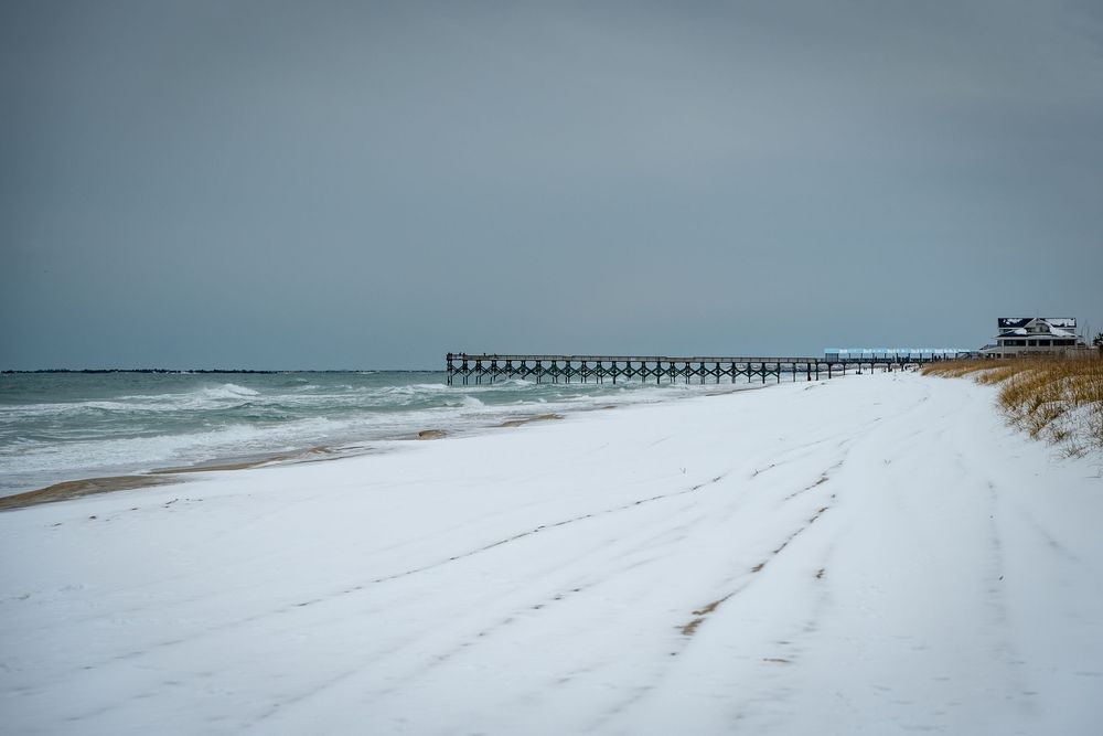 Snow covered beach stretches along the coastline with a wooden pier extending into the gray winter ocean.