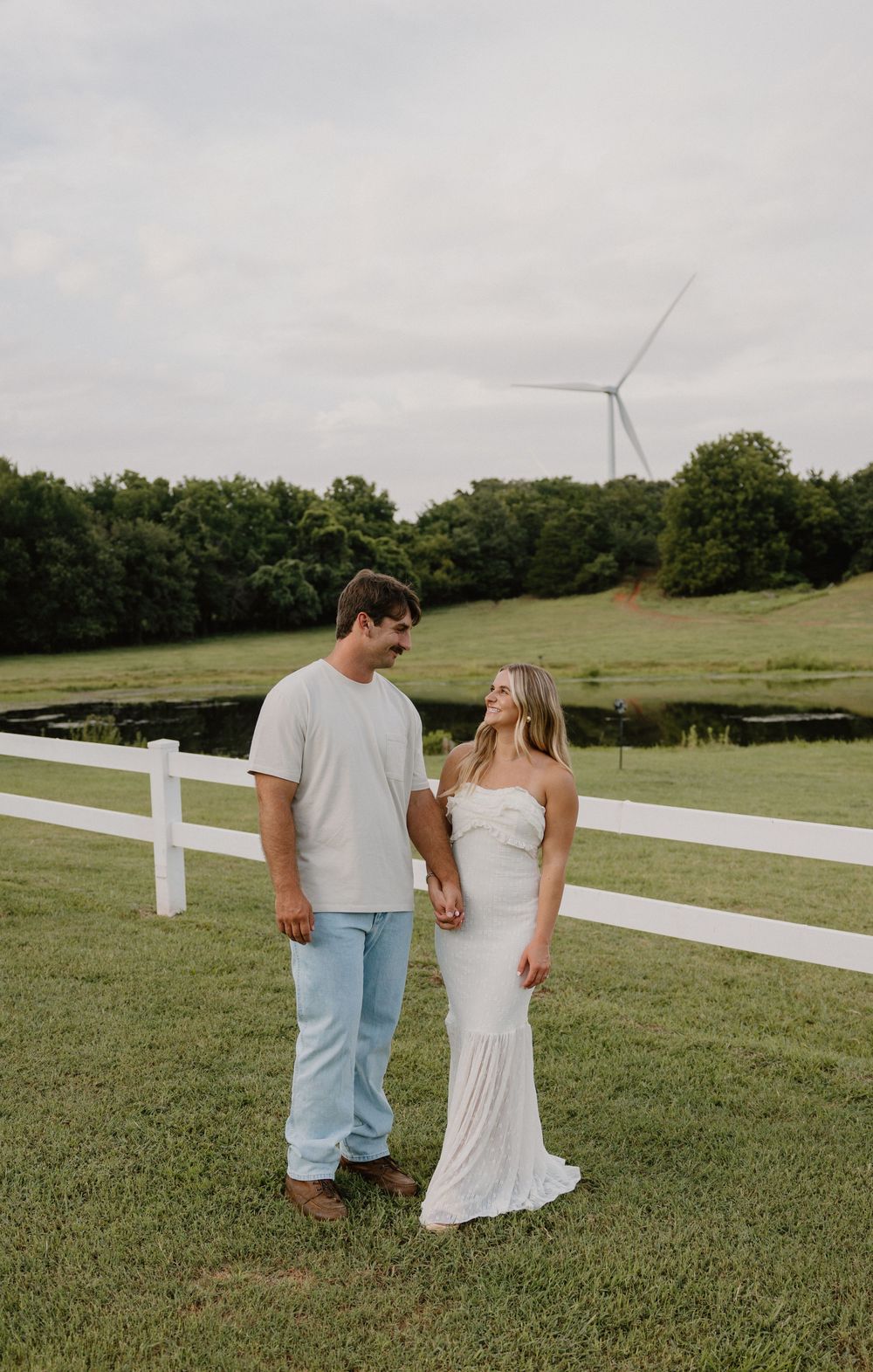 A couple walks together on grassy hills with a white fence and trees in the background.