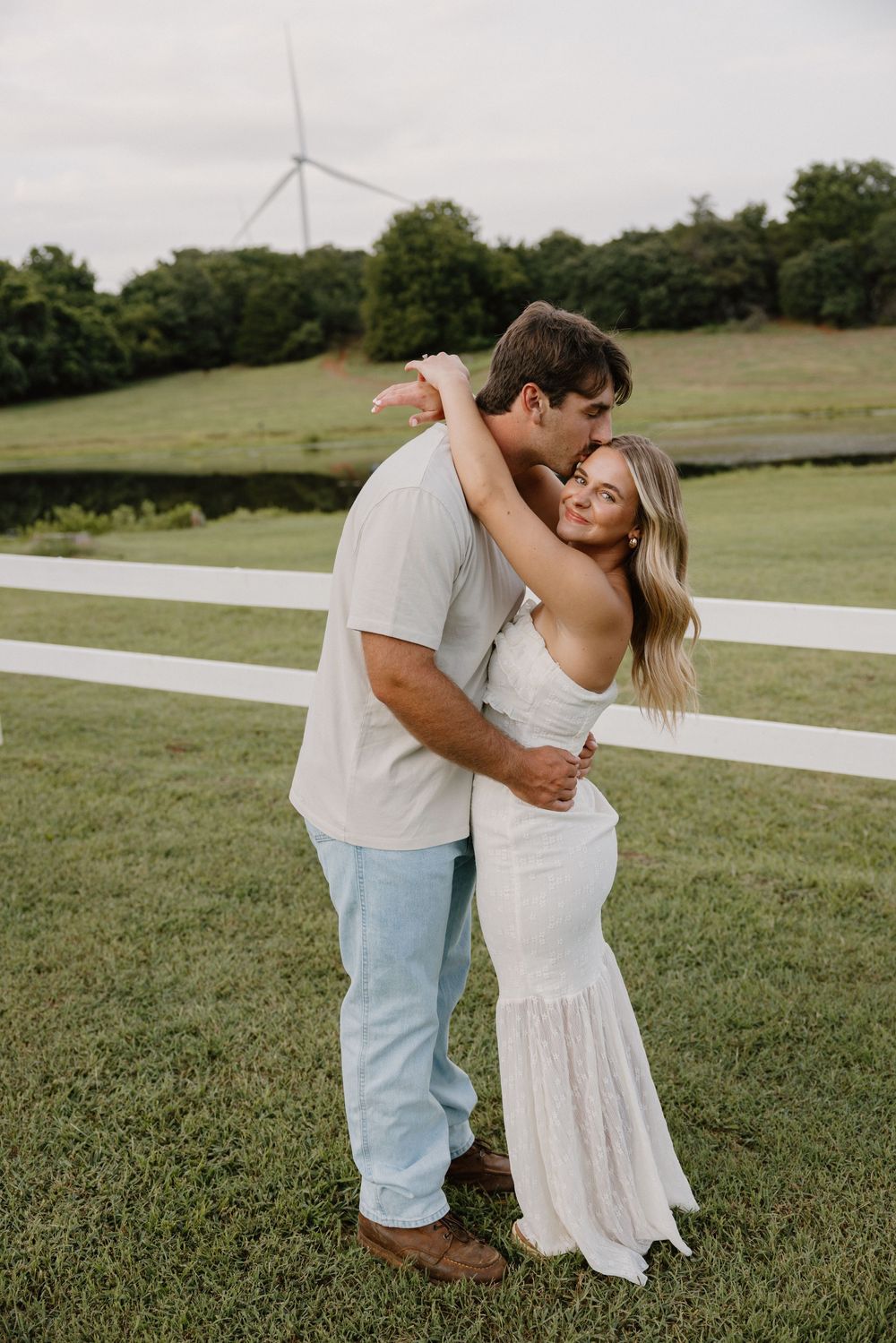 A romantic couple dances together in a grassy field with wind turbines visible in the background.