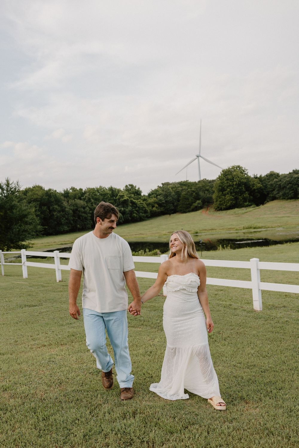 A couple walks hand in hand through a green field with white fencing and wind turbines visible in the background.