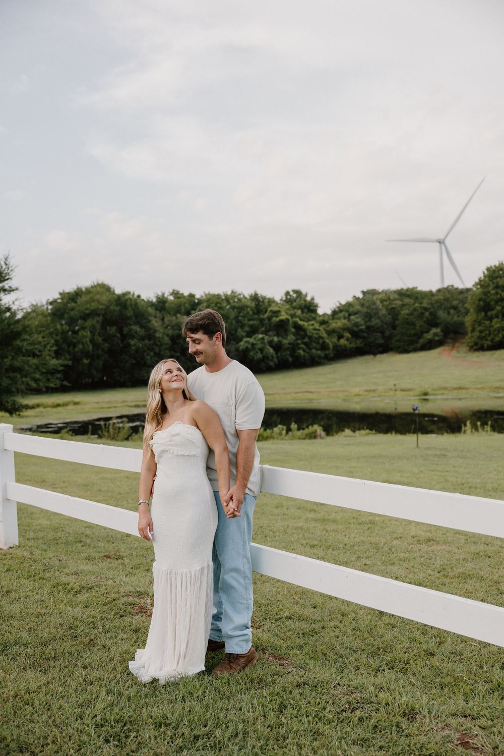 A romantic moment between a couple standing by a white fence on a green hillside.