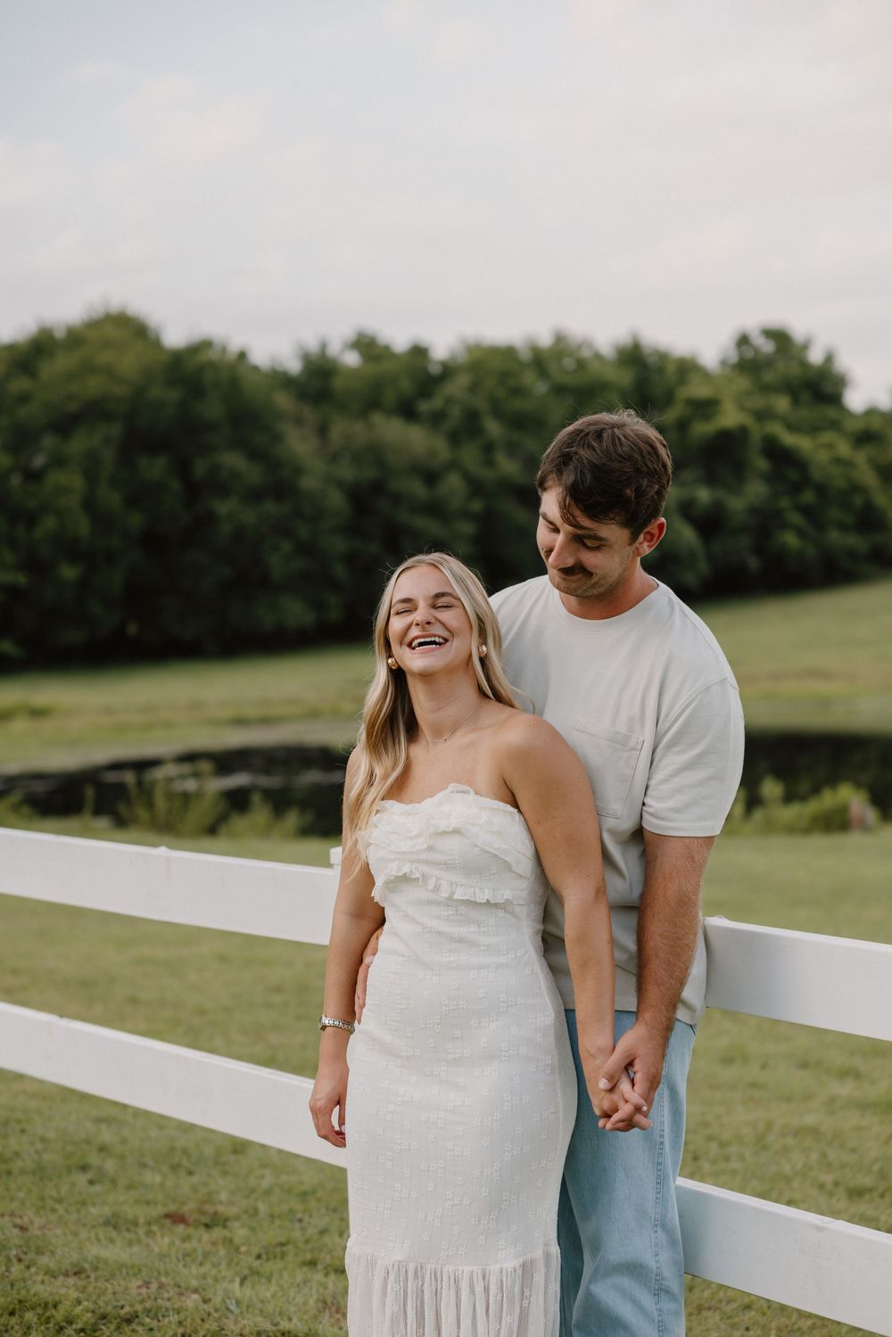 Romantic couple in casual attire walking along white fence in grassy field at sunset.