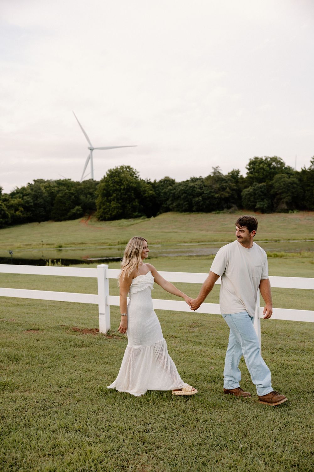 Couple holding hands while walking through pastoral field with white fence and wind turbines.