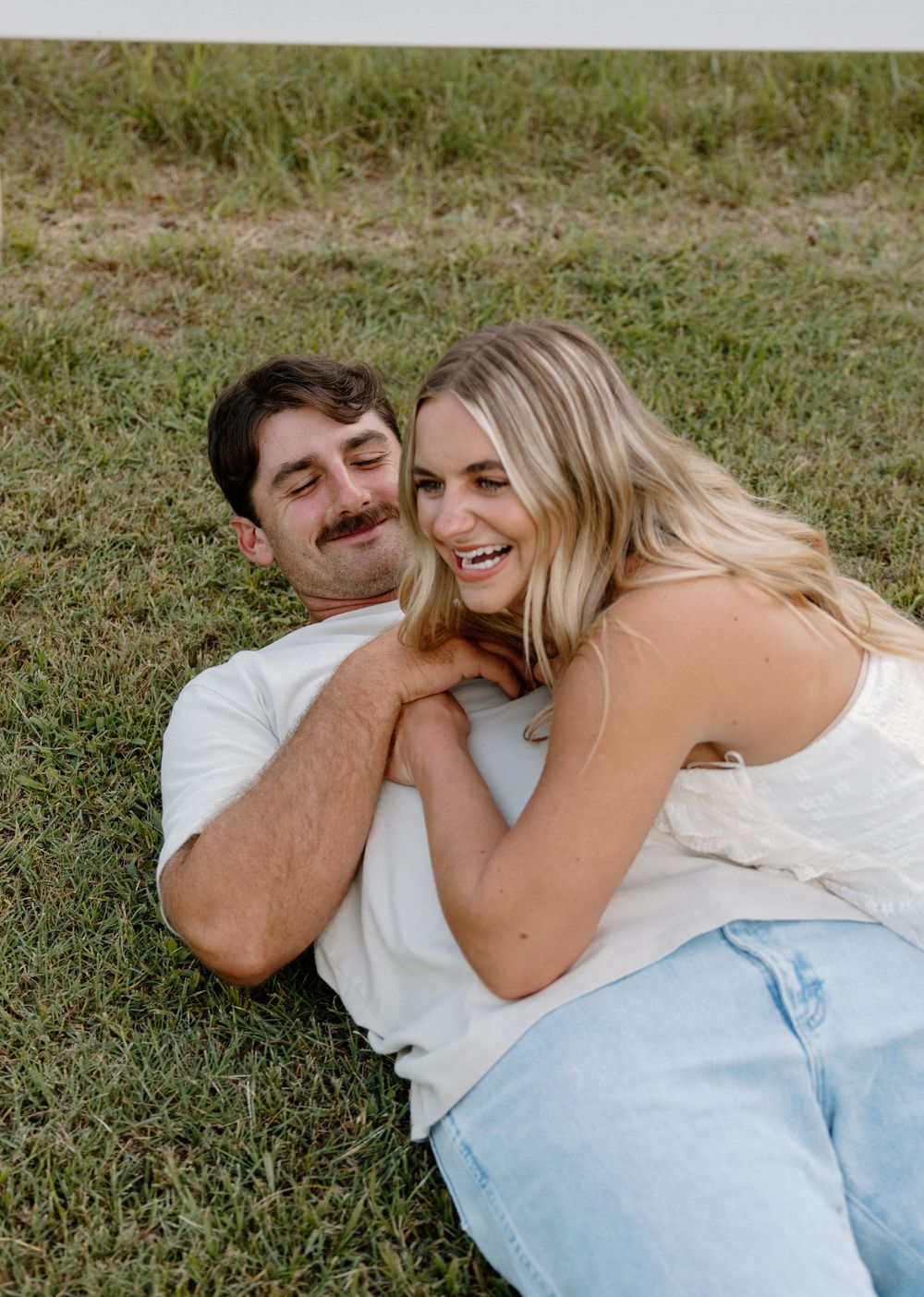 A couple lies together on grass sharing a tender moment in casual white and light blue attire.
