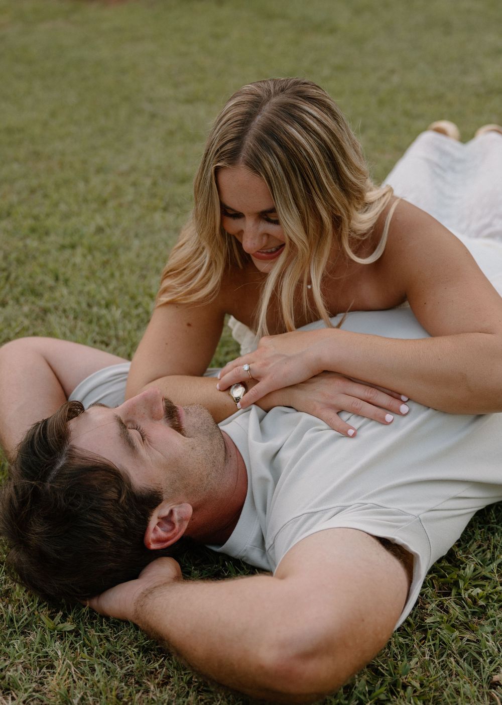 A couple shares playful laughter while lying together on green grass in casual summer clothing.