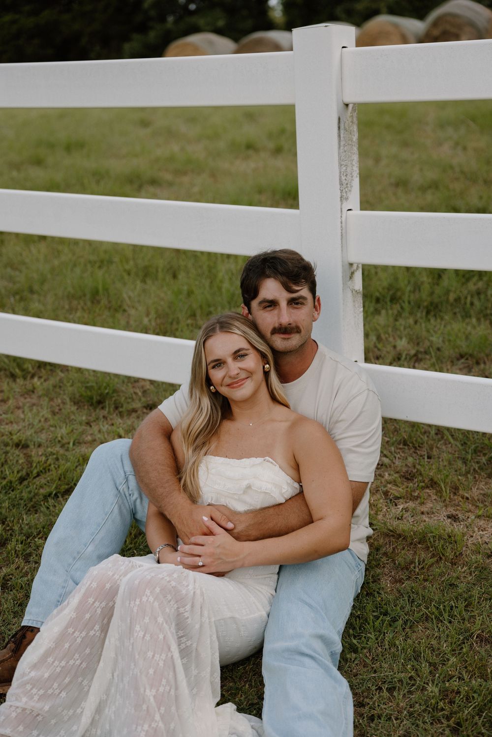 A romantic couple sits together against a white fence in a grassy field during a casual outdoor photoshoot.