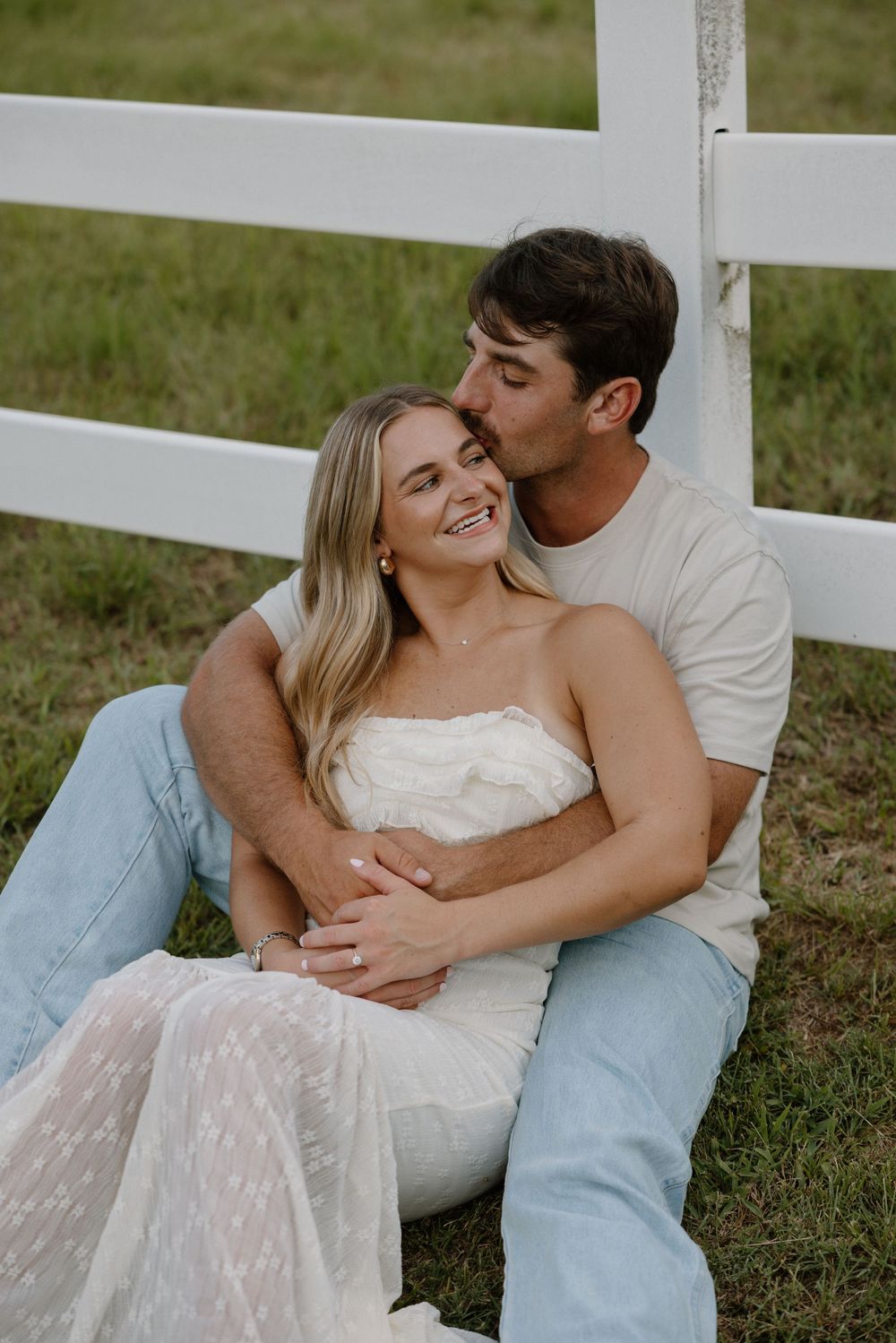 A romantic couple lying together on grass near a white fence, sharing tender moments in casual attire.