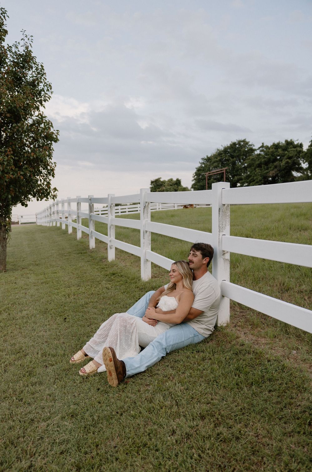 Couple relaxing against white fence in countryside setting during golden hour.