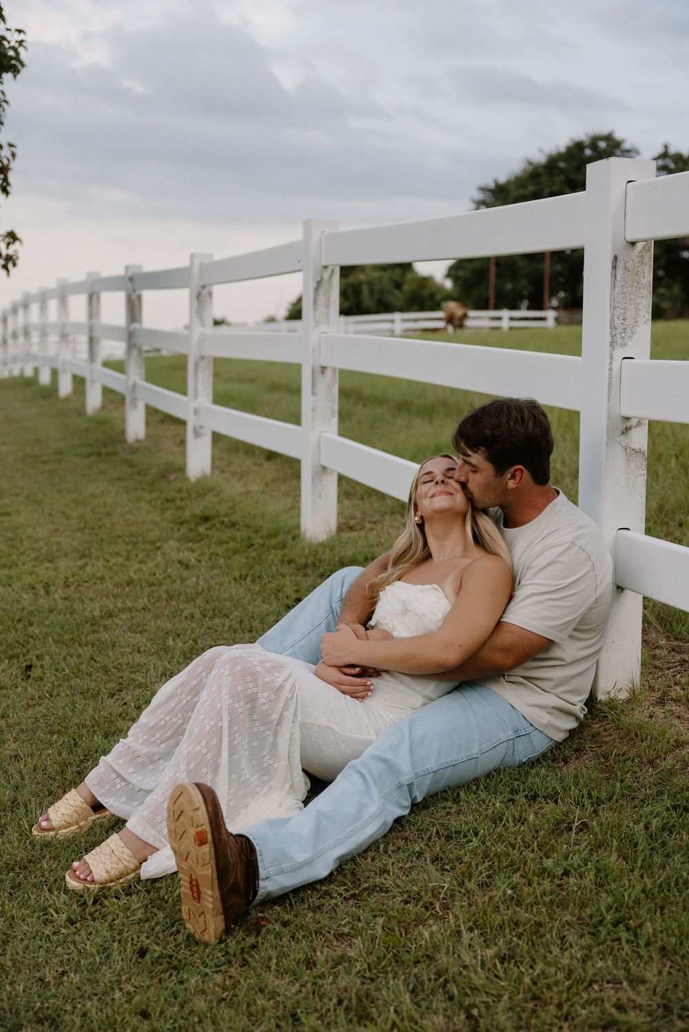 A romantic couple relaxes on grass beside a white fence on a cloudy day, sharing an intimate moment together.