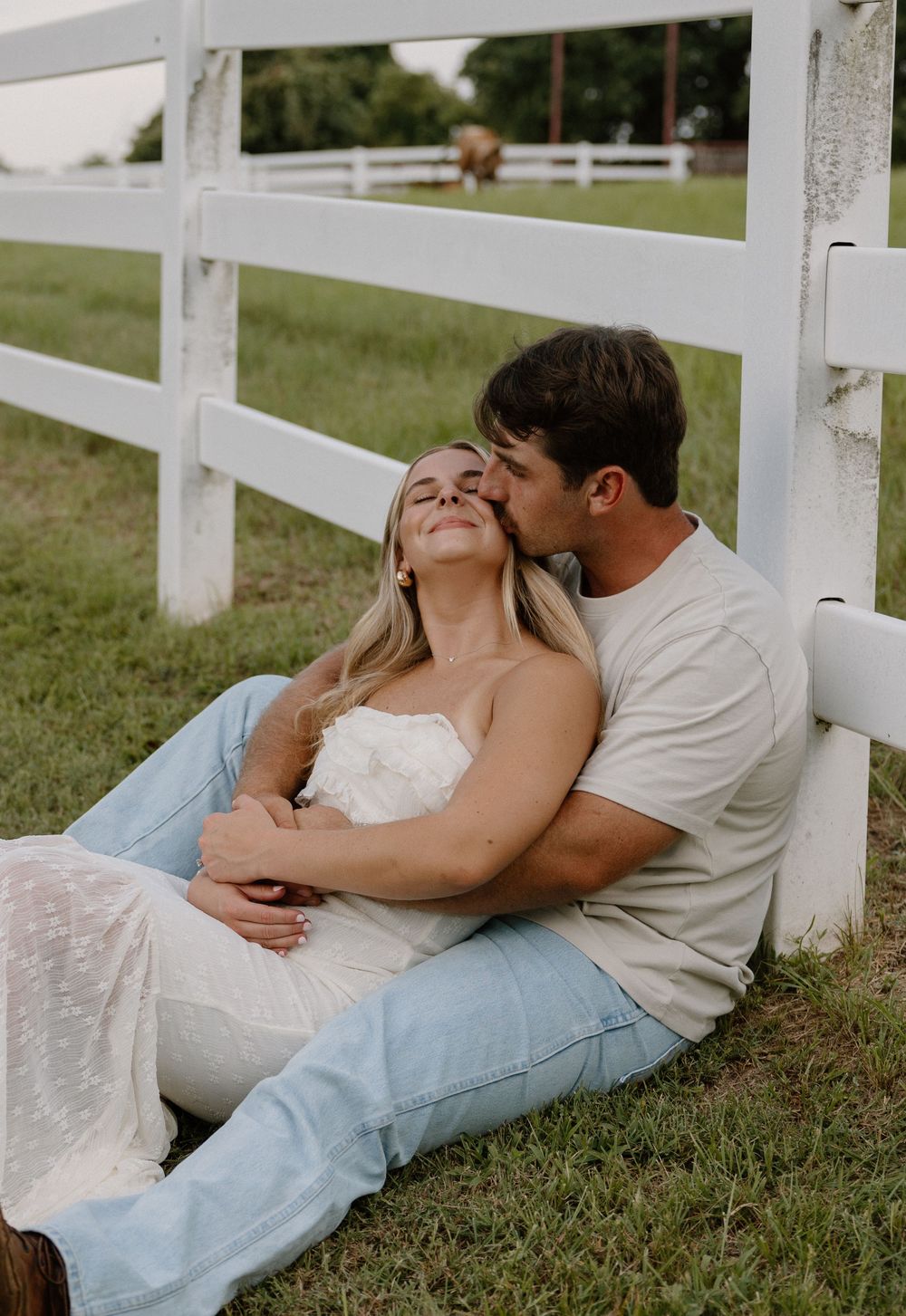 A romantic couple in casual attire shares tender moments while lying together along a white fence on grassy ground.