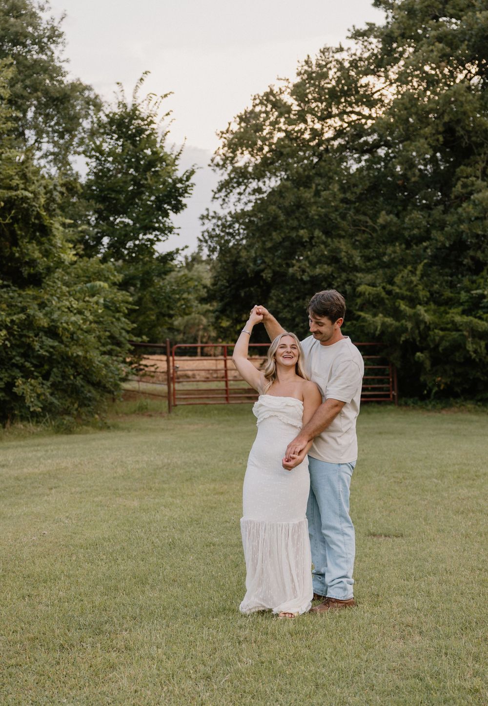 A couple dances together in a grassy field with trees and a red gate in the background.