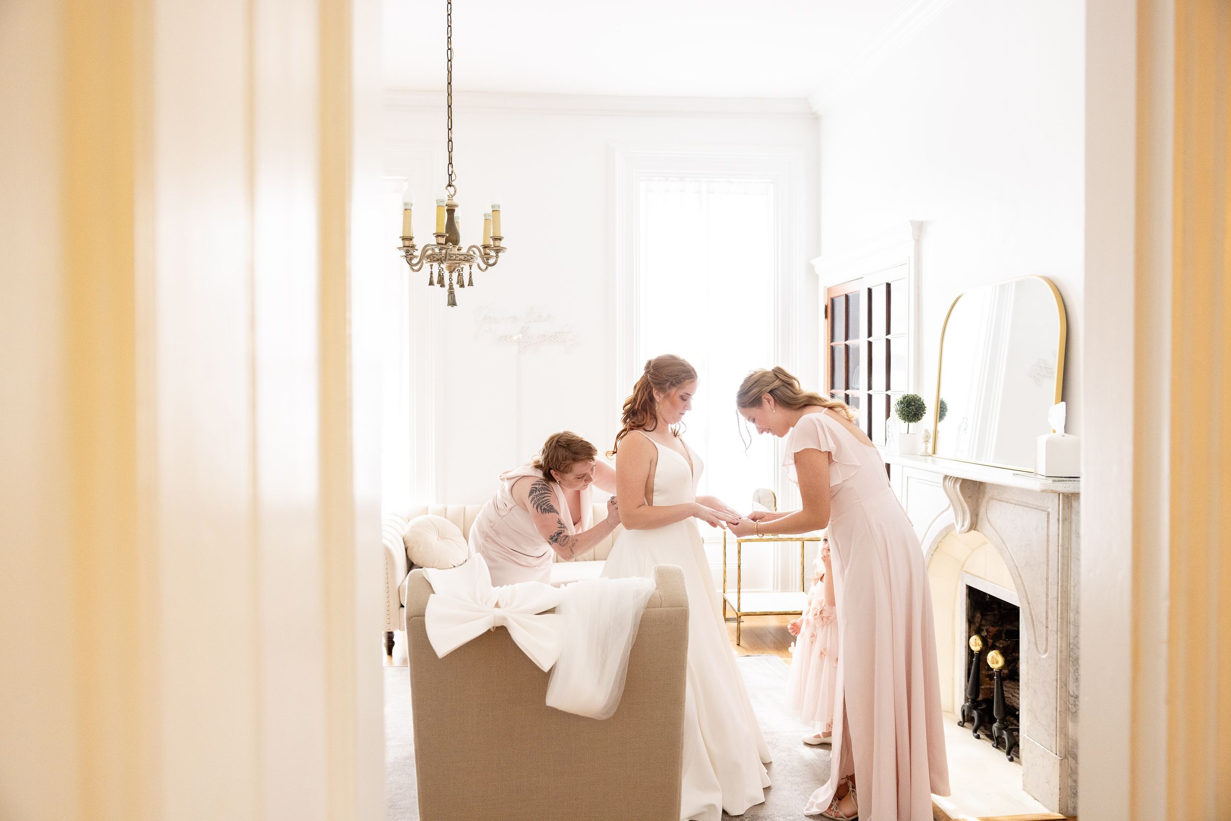 A bride and bridesmaids getting ready for a wedding in a bright, airy room with a chandelier.