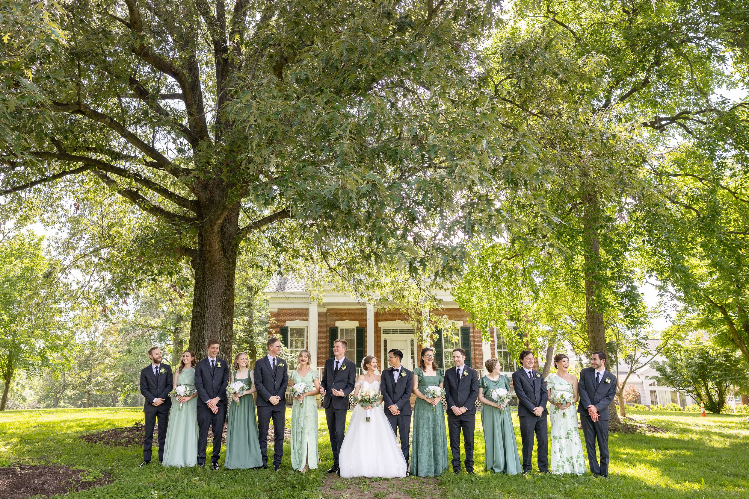 A wedding party posing outdoors in a lush, green park setting with tall trees providing shade.