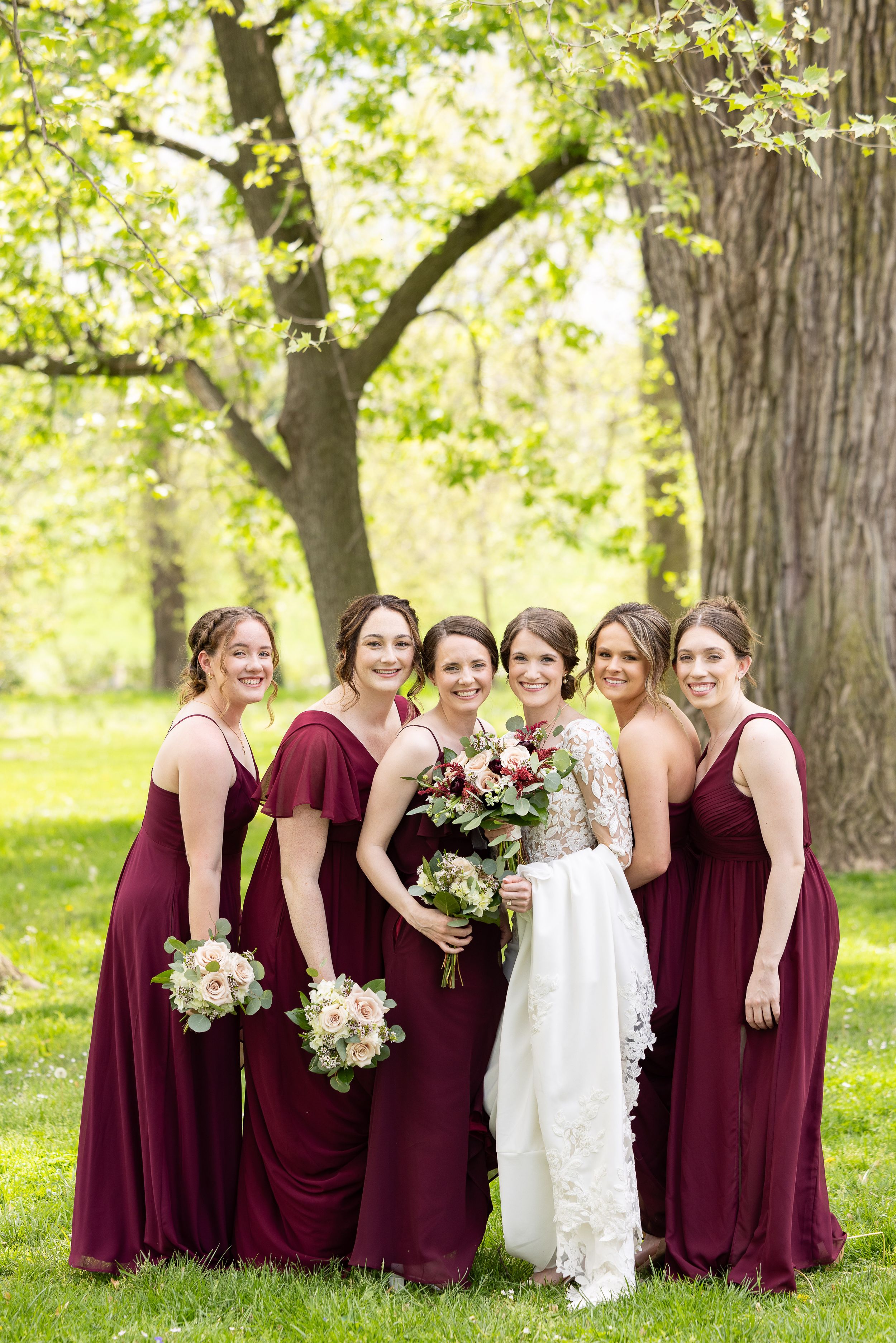 A bride and her bridesmaids in burgundy dresses standing together in a lush green park.