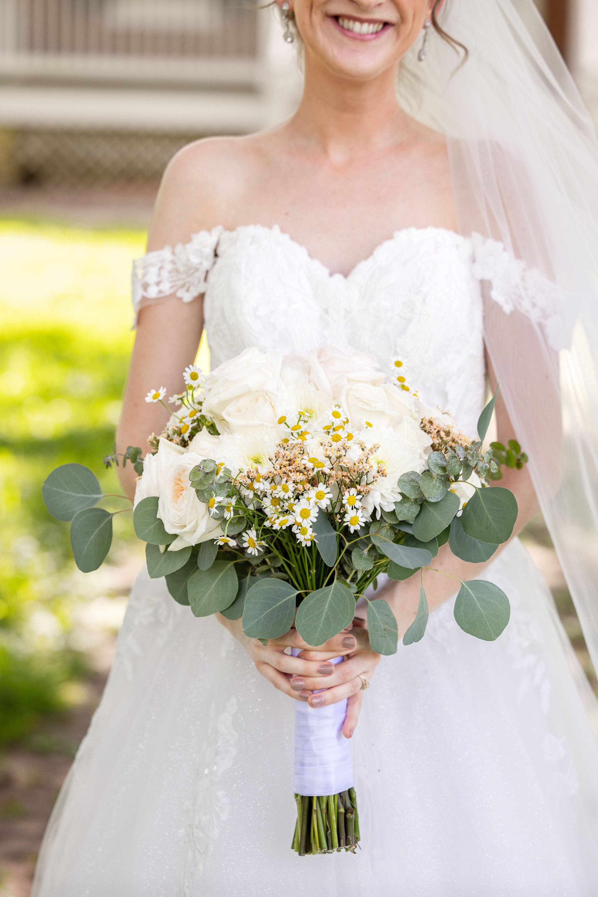 A smiling bride in a white off-the-shoulder wedding dress holding a bouquet of flowers in a garden setting.