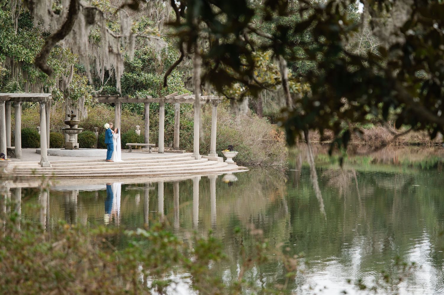 Couple embracing on a stone colonnade gazebo reflected in a tranquil pond surrounded by moss-draped trees.