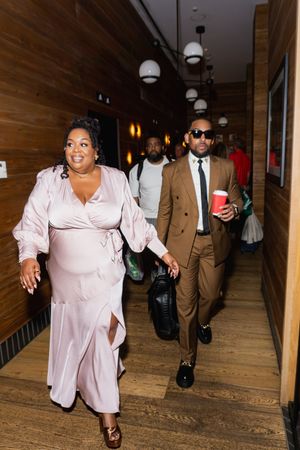 Stylish couple walking in hallway, woman in pink satin dress, man in brown suit holding red cup.
