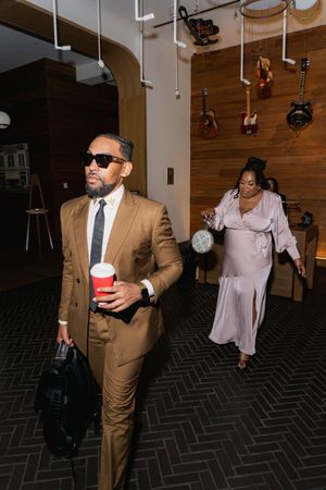 Man in tan suit with sunglasses holds red cup while woman in pink gown walks through music-themed venue.