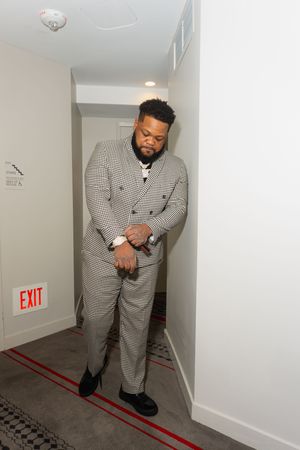 Man in gray houndstooth double-breasted suit standing in hallway near exit sign, looking down at his hands.
