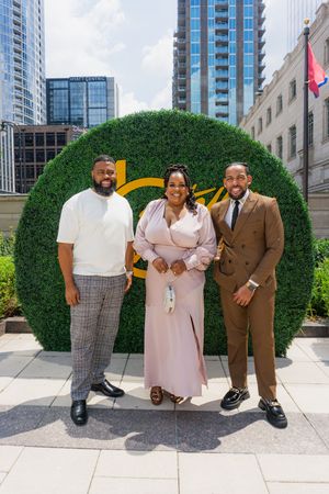 Three well-dressed individuals posing in front of a decorative green hedge wall in an urban setting.