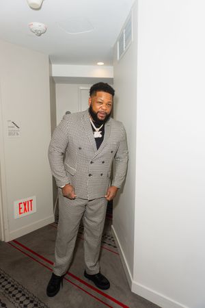 Man in gray houndstooth double-breasted suit with chain necklace standing near exit sign in hallway.