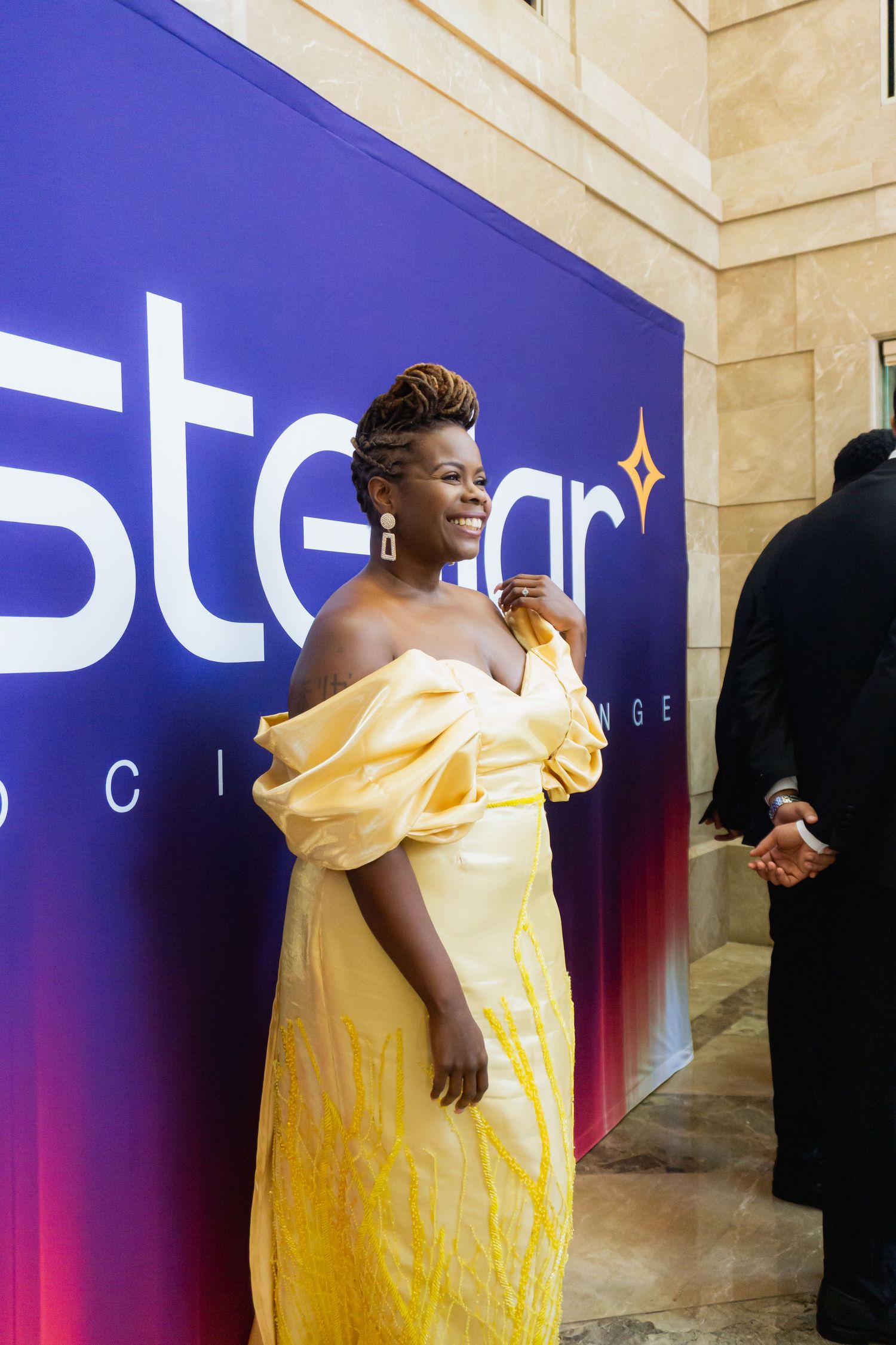 Woman in elegant yellow off-shoulder gown smiling at Stellar awards event on a purple branded backdrop.