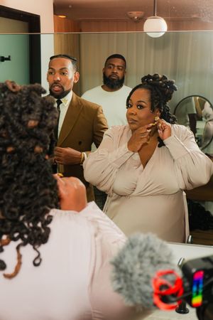 Woman putting on earrings while getting ready, reflected in mirror with two men in formal attire behind her.