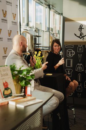 Two people sit and converse with microphones at a Vanderbilt Bookstore event with books and flowers nearby.