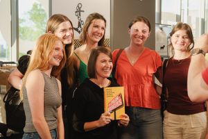 A smiling woman holds a yellow book surrounded by six young women at a casual indoor gathering.