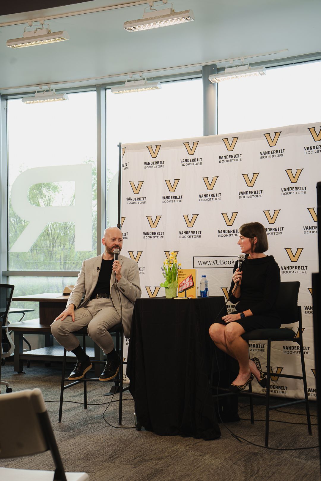 Two speakers hold microphones at a Vanderbilt Bookstore event, seated in chairs before a branded step-and-repeat backdrop.