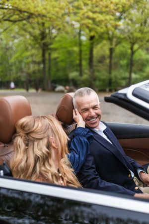 A couple shares a joyful moment together in a luxury convertible car with trees in the background.
