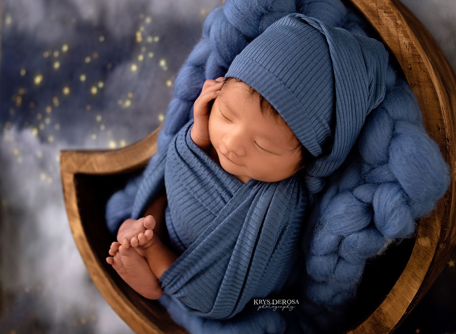 Sleeping newborn wrapped in blue textured fabric in wooden bowl against starry backdrop.