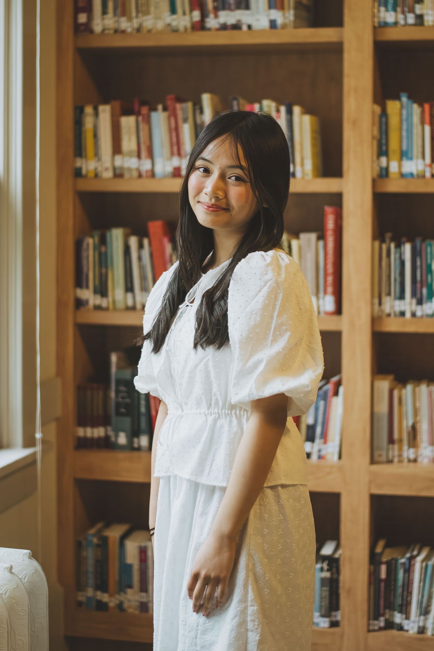 A student in a white dress stands in a library surrounded by wooden bookshelves filled with books.