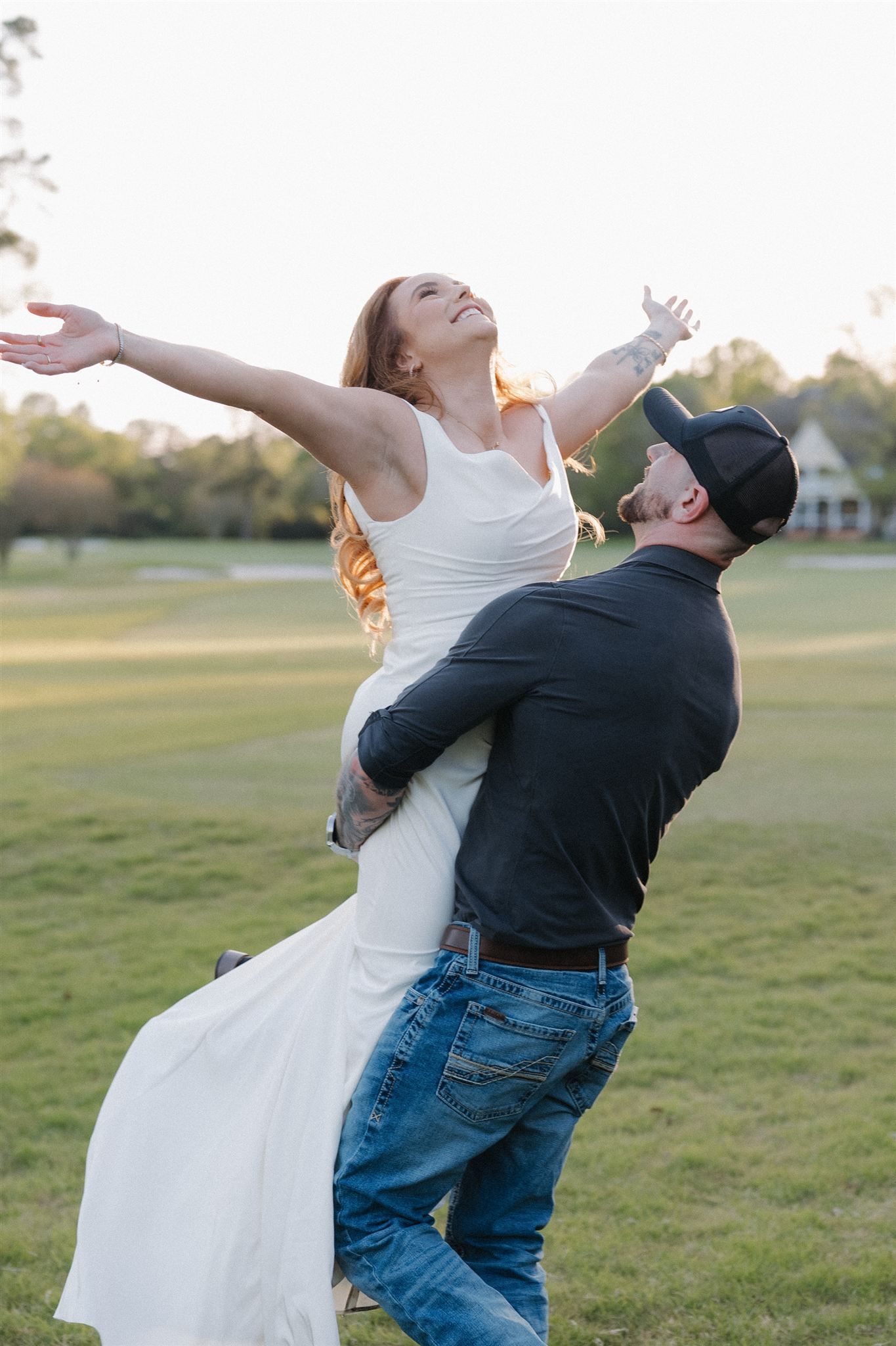 A couple in wedding attire performs a playful dip pose together on a grassy field at sunset.