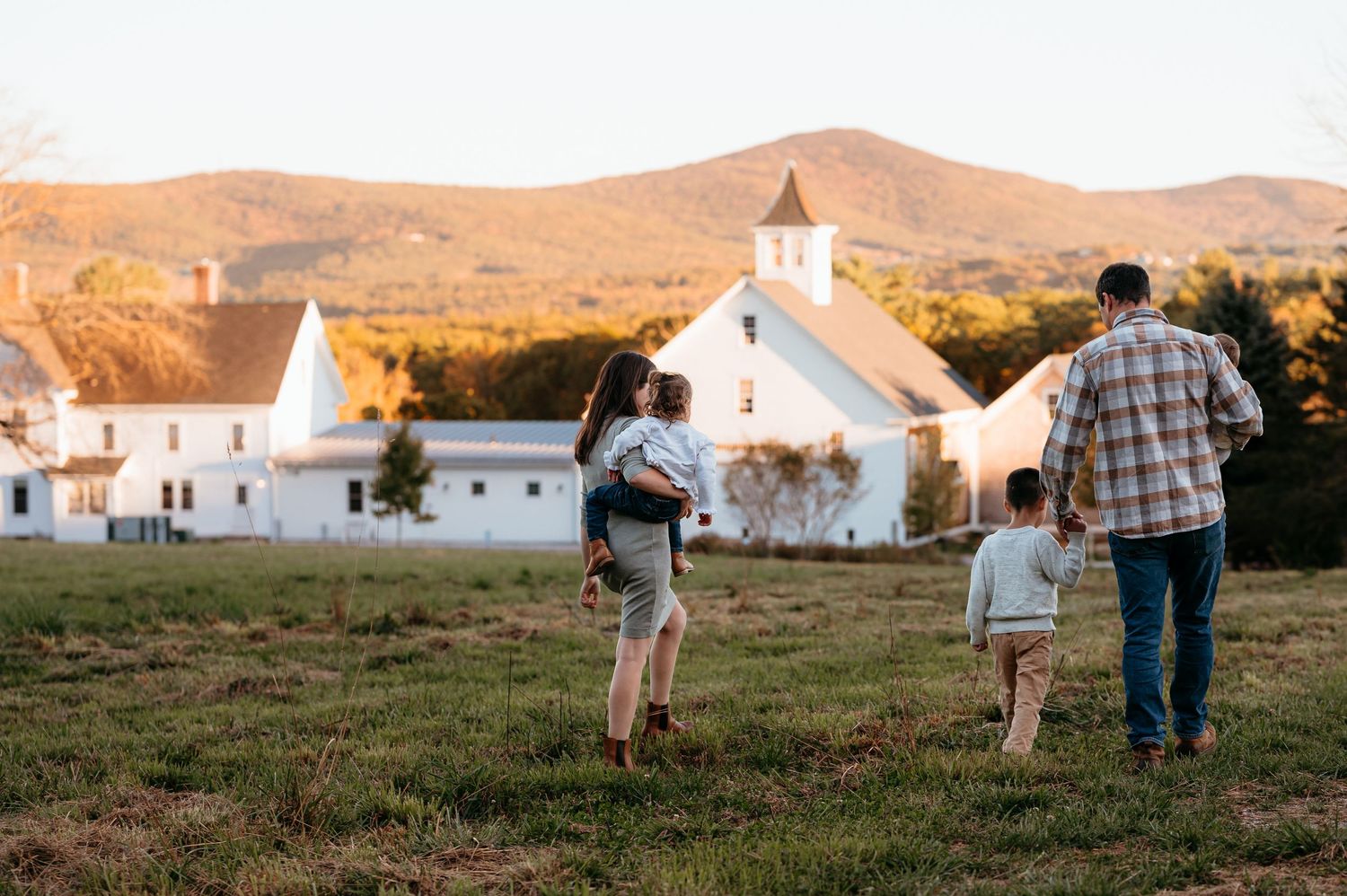 Family walking toward white farmhouse and church with mountain backdrop at golden hour sunset.