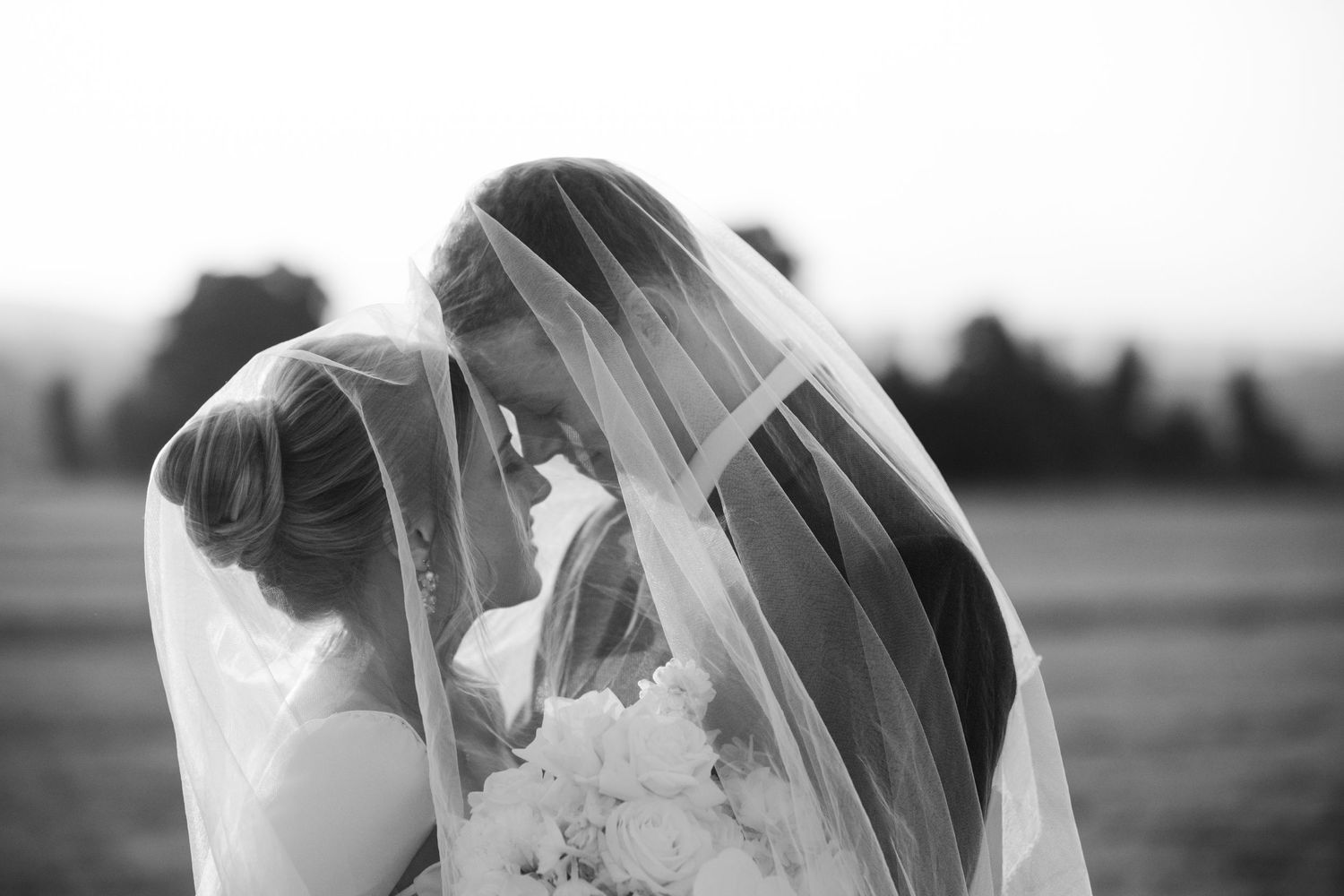 Romantic black and white photo of a bride and groom sharing a kiss beneath a flowing wedding veil.