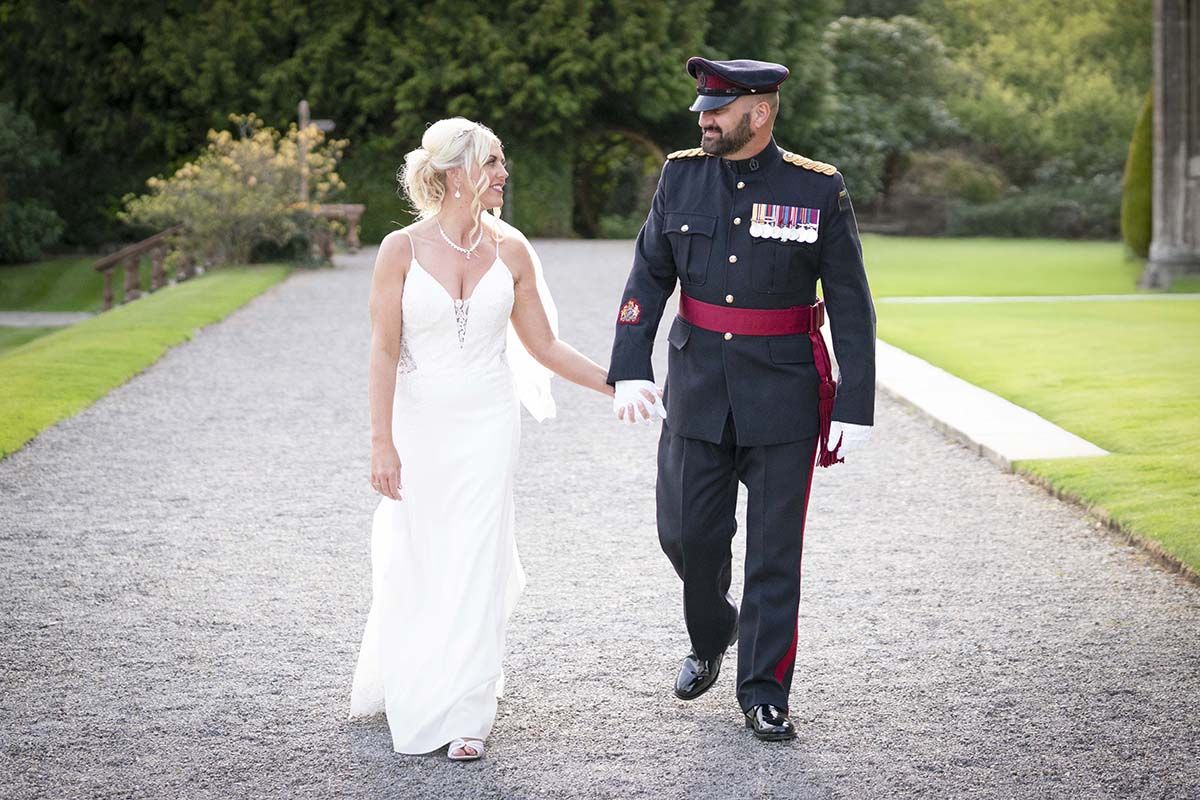 A couple walks together on a gravel path, with the bride in white and groom in military dress uniform.