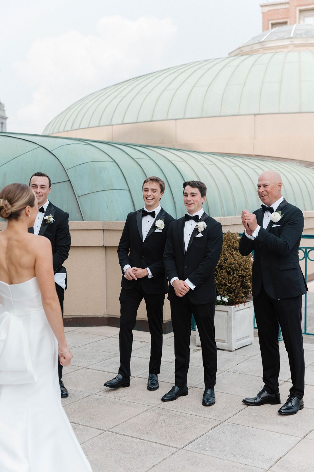 Groomsmen in black tuxedos line up for a playful wedding photo on a rooftop with domed architectural features.