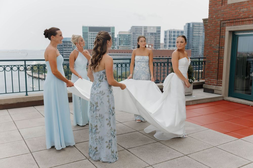 Bridesmaids in pastel dresses help arrange a wedding gown on an urban rooftop terrace with city skyline views.