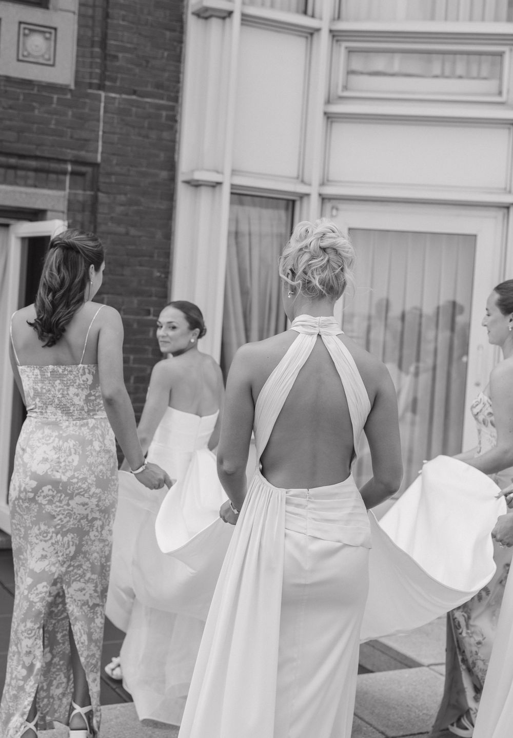 A group of women in floral dresses and white aprons stand in a line looking through windows of a brick building.
