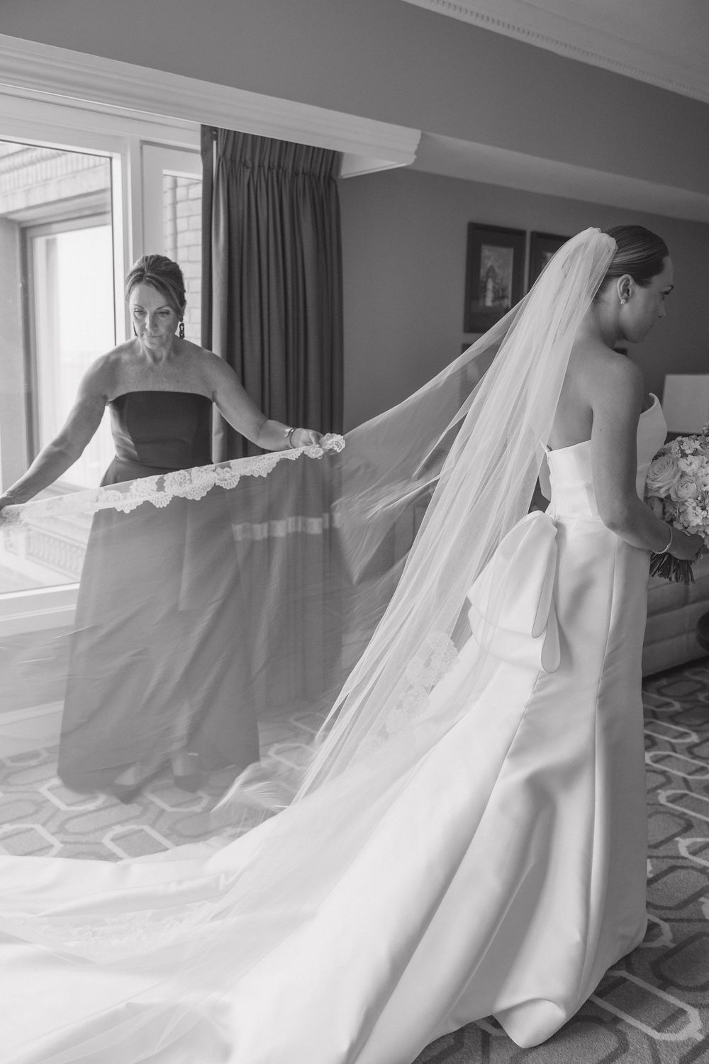 A flowing cathedral-length veil trails behind a white wedding dress as it moves gracefully across a carpeted hotel room floor.