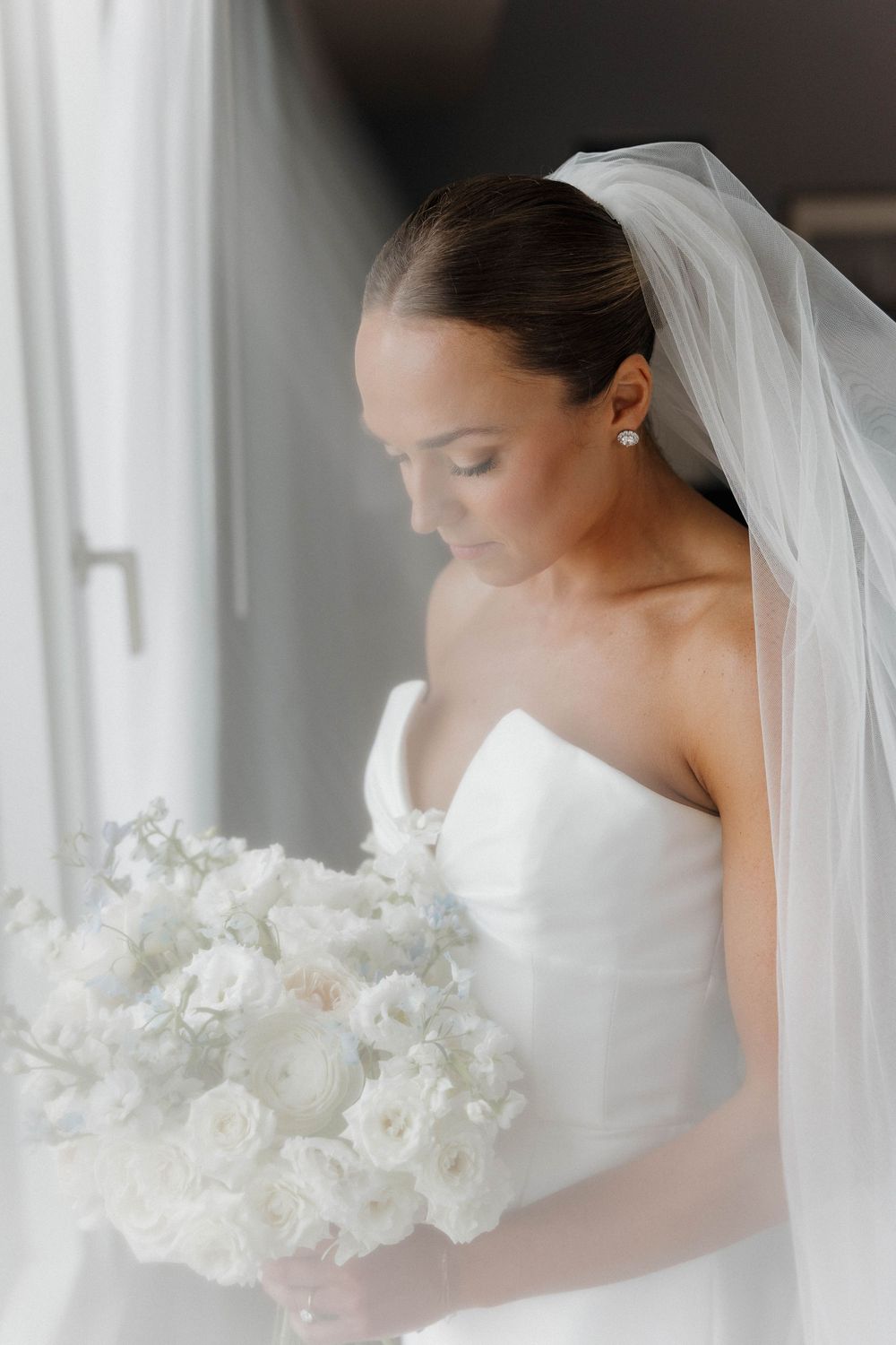 A bride in a white wedding dress and veil holds an elegant bouquet of white flowers in a soft, dreamy setting.