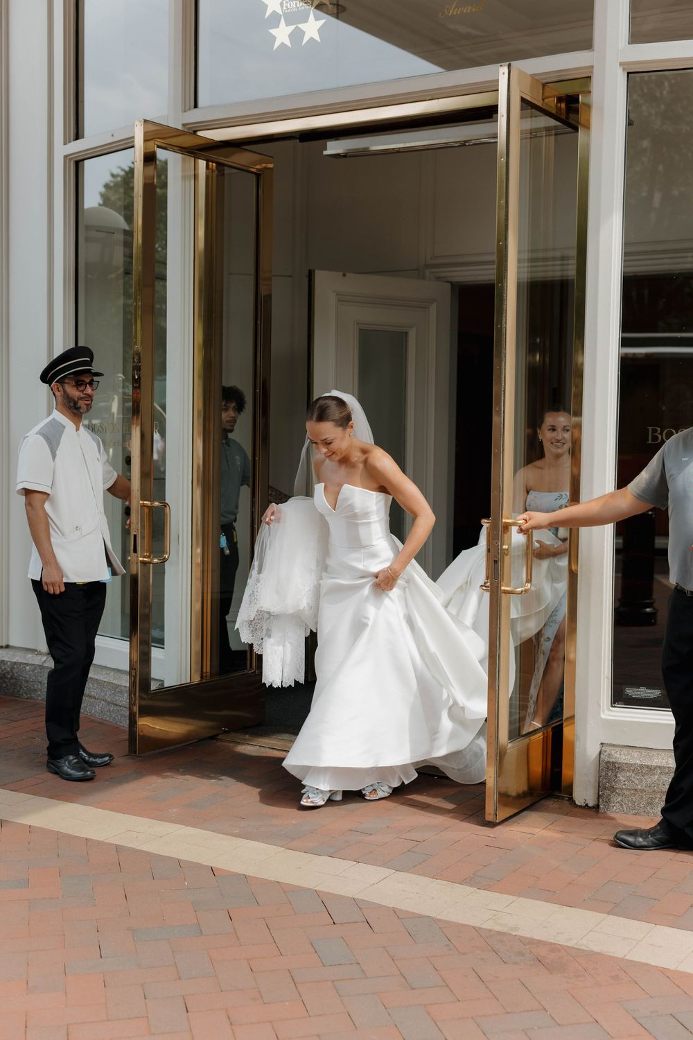 A bride in a white wedding dress spins joyfully in a hallway with brick flooring while others watch from doorways.