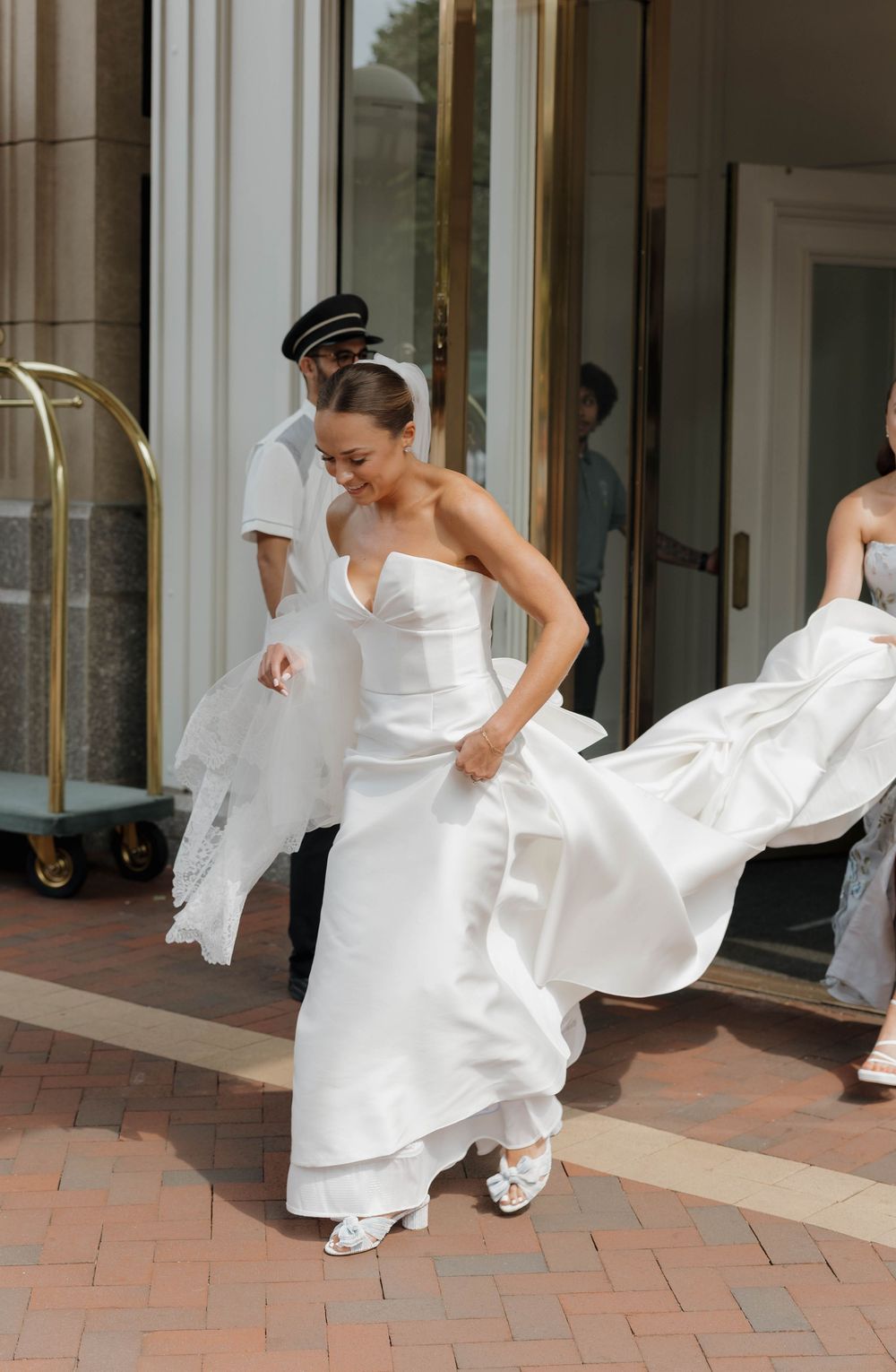 A bride in a flowing white wedding dress twirls and dances on a brick patio outside a building.
